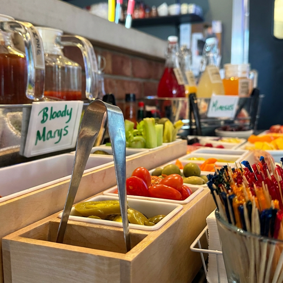 Bloody Mary bar with pitchers of tomato mix, vegetable garnishes, condiments, tongs, and colorful cocktail stirrers.