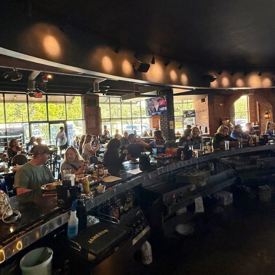 Busy restaurant interior with diners seated at tables and a bar, warm lighting, large windows, and a pink neon sign.