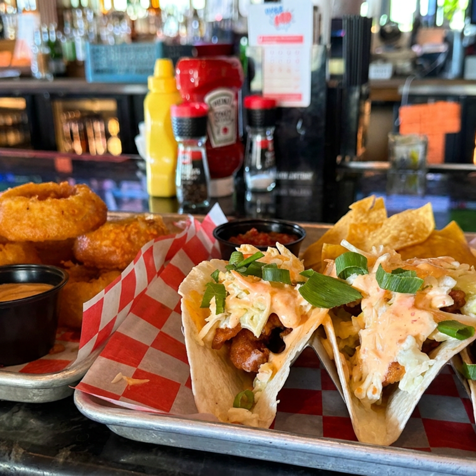 Crispy fish tacos with slaw and green onions beside golden fritters, dipping sauces, condiments, and a cocktail at a bar.