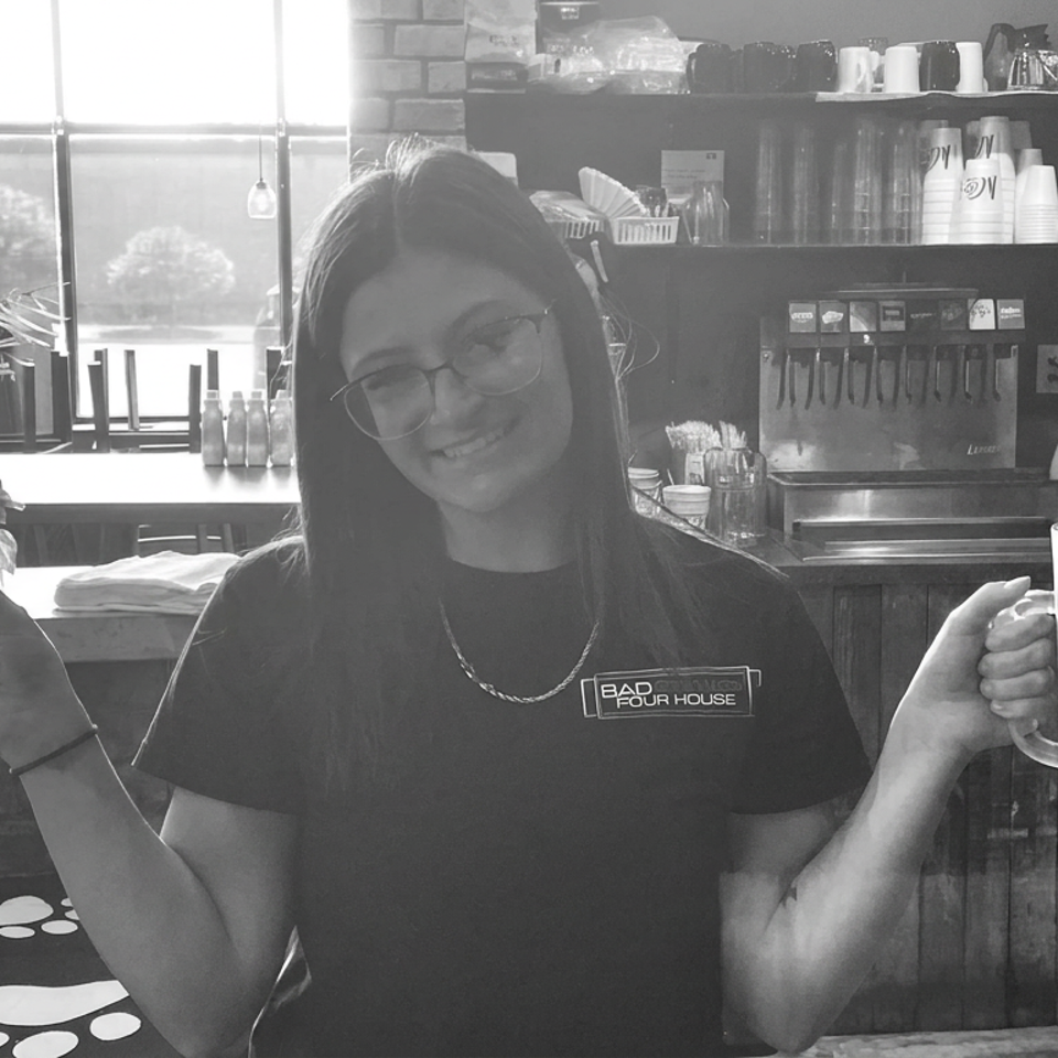Smiling restaurant employee holds two large frosted beer mugs inside a casual bar with beverage taps and shelves.