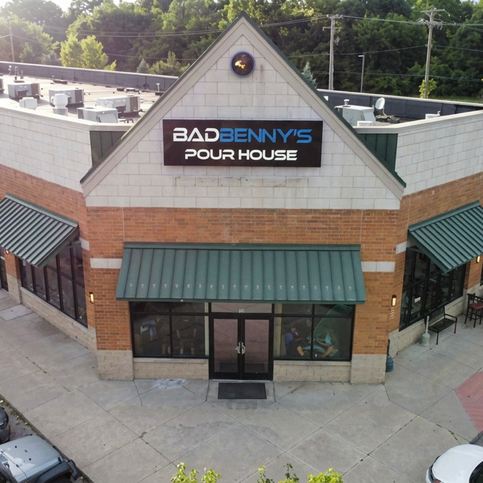 Aerial view of Bad Benny’s Pour House restaurant exterior with green awnings, outdoor patio seating, parked cars, and wooded surroundings.